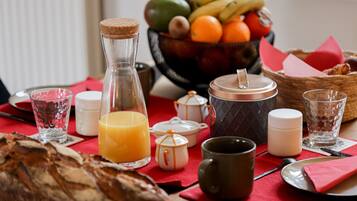 A breakfast setting with bread, a fruit bowl, a bottle of orange juice, and various breakfast items on a red tablecloth.