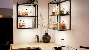 Una cocina con dos estantes negros que sostienen varias botellas y frascos, un florero negro con ramas y una tetera sobre la encimera.