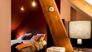 Un dormitorio con techo inclinado, una cama con varias almohadas, una lámpara de noche y una mesita con una caja.