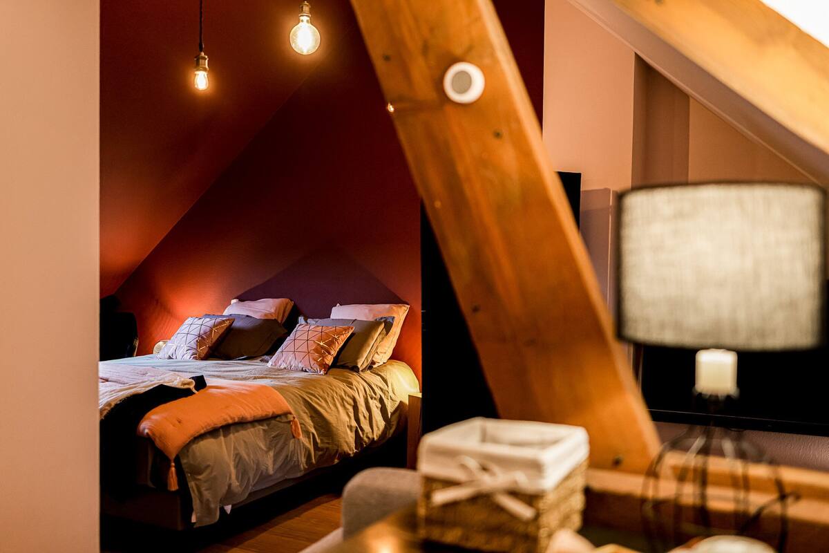 A bedroom with a sloped ceiling, a bed with multiple pillows, a bedside lamp, and a small table with a box.