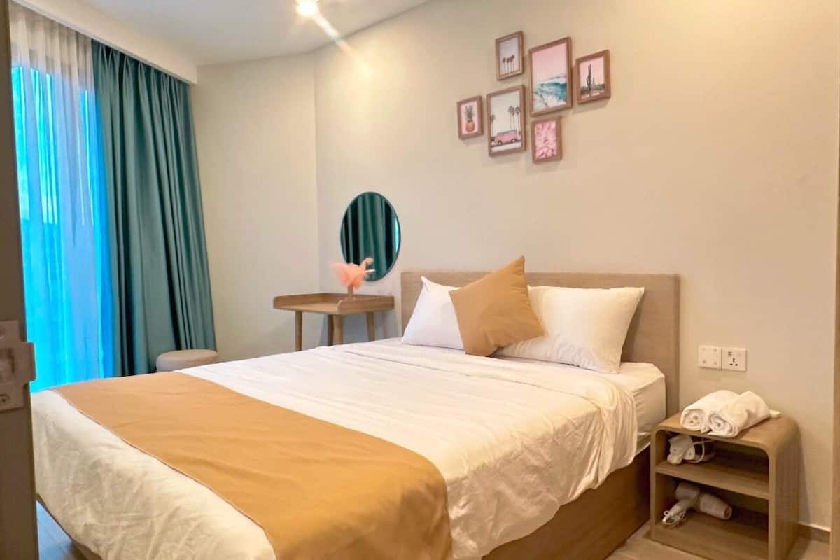 A hotel room with a large bed, a bedside table with a mirror, a wall-mounted shelf with towels, and a window with curtains.