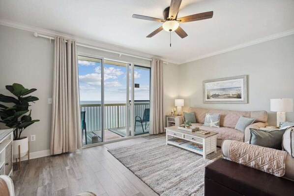 A spacious living room with a large sliding glass door leading to a balcony, a ceiling fan, a sofa set, and a coffee table.