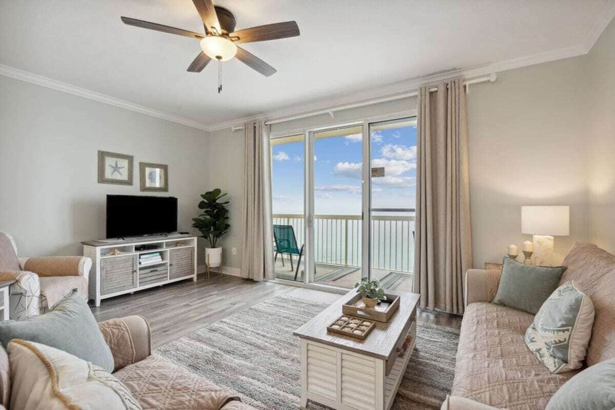A spacious living room with a ceiling fan, a flat-screen TV, a coffee table, and a sofa. There is a sliding glass door leading to a balcony with a view of the sky.
