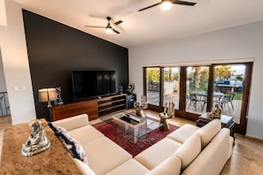 Un salon moderne dotĂ© dâun tĂ©lĂ©viseur Ă Ă©cran plat, dâun ventilateur de plafond, dâune grande fenĂȘtre et dâun coin repas avec des siĂšges extĂ©rieurs.