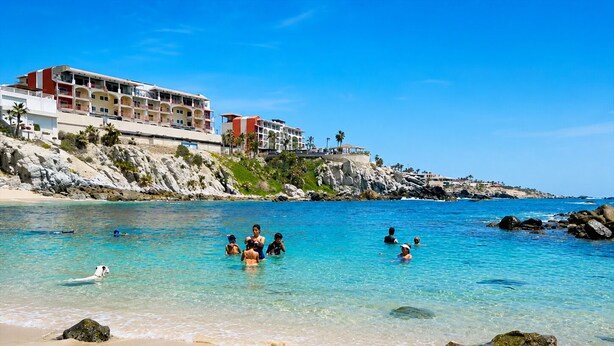 Una platja d'aigua cristal·lina, persones nedant i un hotel al fons amb nombrosos pisos i balcons.