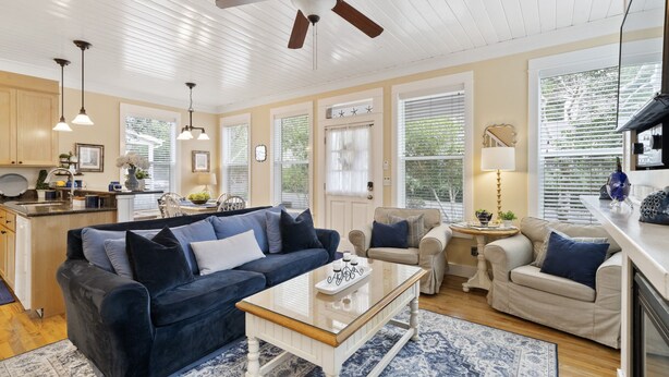 Un salon spacieux avec un canapé bleu foncé, une table basse blanche et un fauteuil beige. On y trouve un ventilateur de plafond, un coin repas avec une table et des chaises, ainsi que de grandes fenêtres équipées de stores.