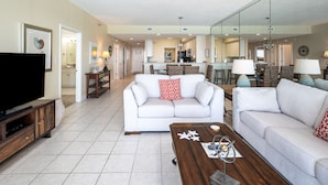 A spacious living room with a large flat-screen TV, two white sofas, a wooden coffee table, and a kitchen in the background. -  "Miles-of-Smiles"- Navarre Beach Regency #611 (Navarre)