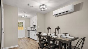 Un comedor con una mesa de madera preparada para cuatro, una cocina al fondo y un equipo de aire acondicionado en la pared.