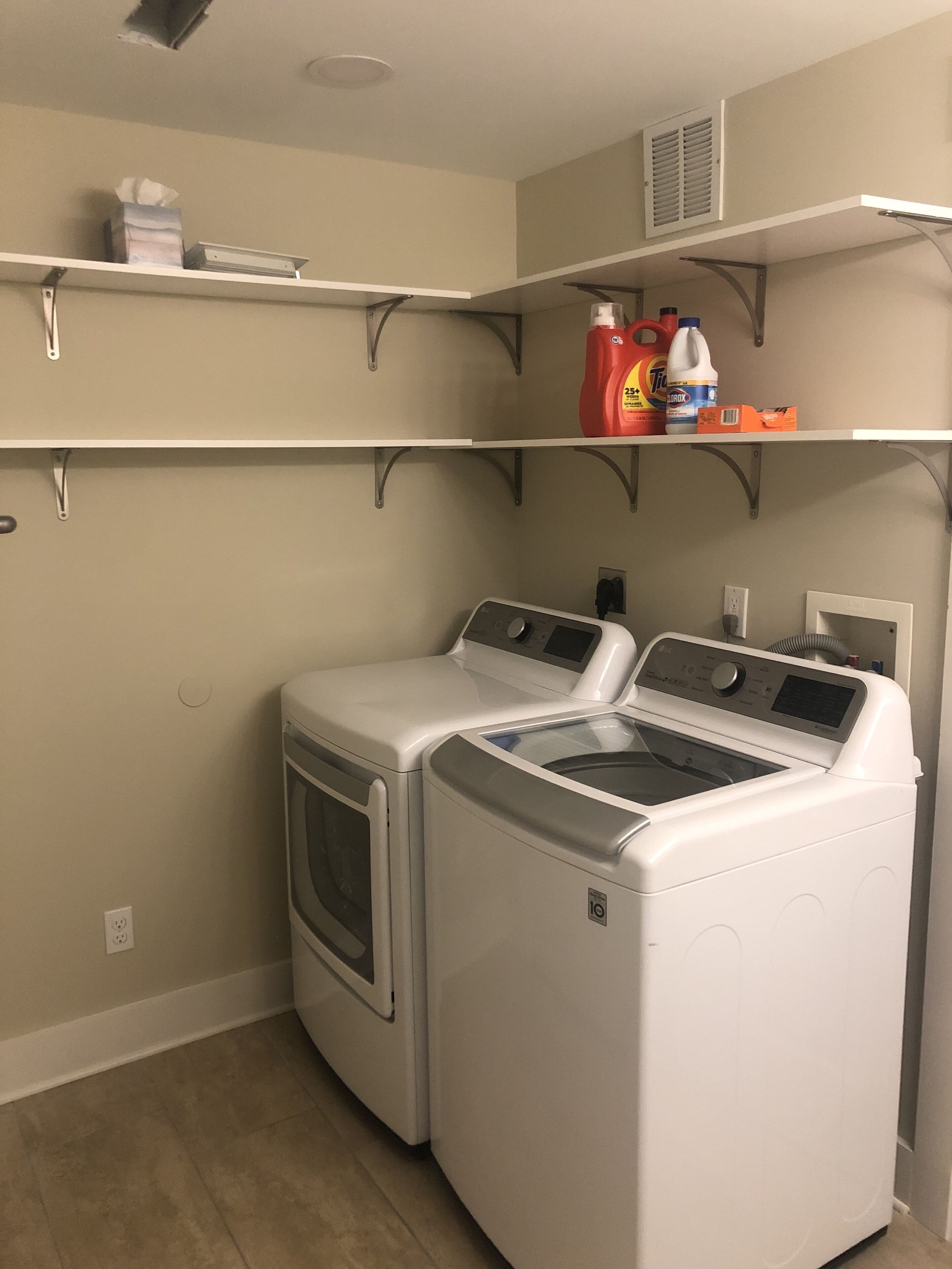 A laundry room with two front-loading washers and dryers, a shelf with cleaning supplies, and a vent on the wall.