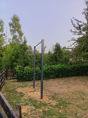 Un espace extérieur doté d’une structure métallique, peut-être une structure d’escalade, entouré de verdure et délimité par une clôture en bois.