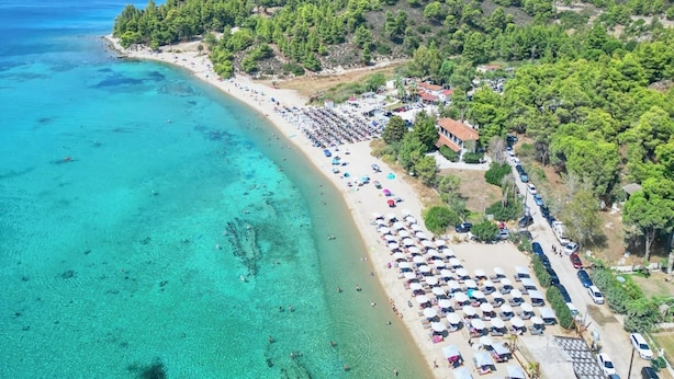 Uma área costeira com praia, águas turquesa cristalinas e uma fileira de carros estacionados à beira-mar.