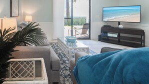 A living room with a sofa, a coffee table, and a television. There is a view of the outdoors through a glass door.