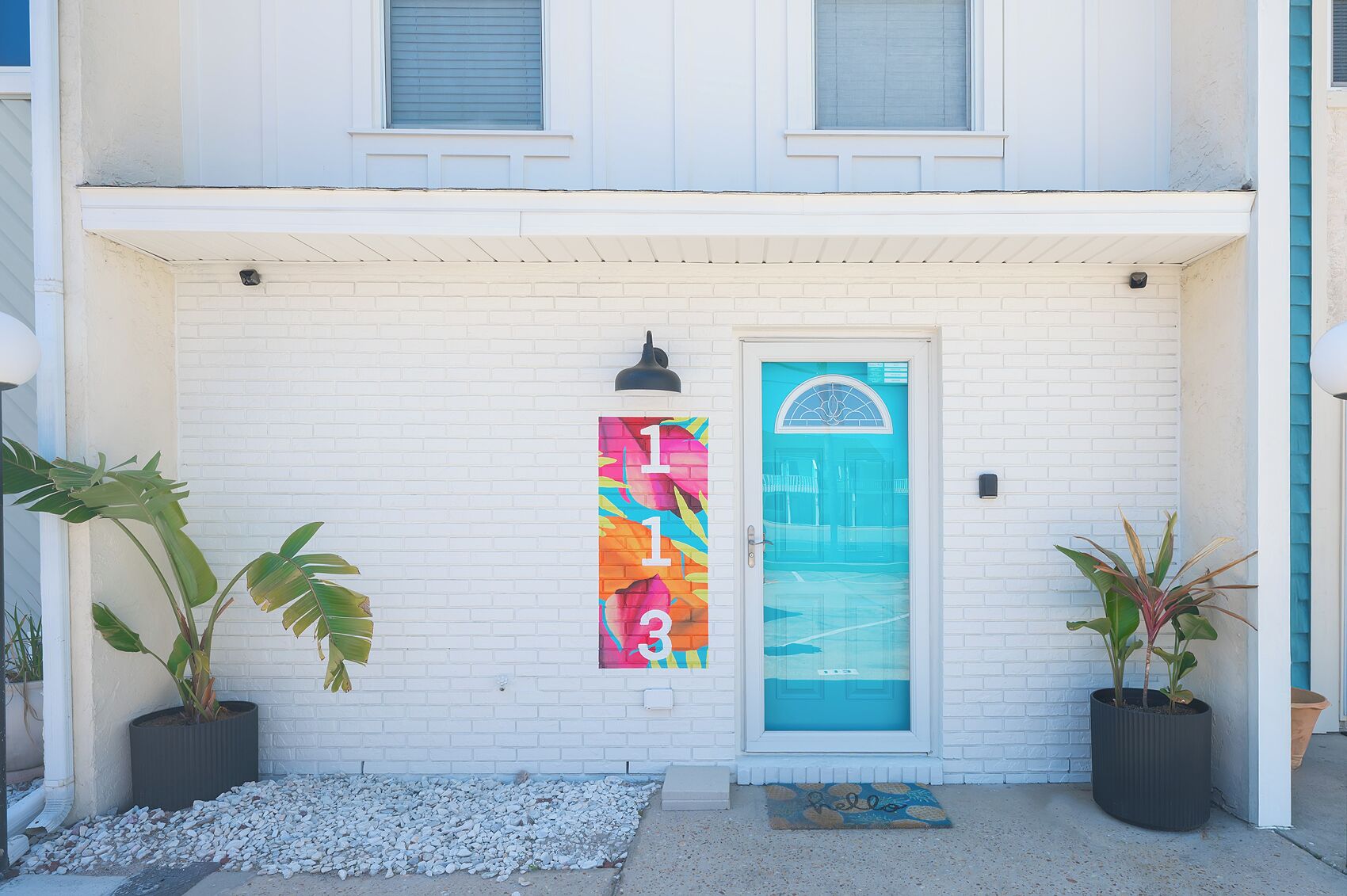 A white brick building with a blue door, a colorful vertical sign, and two potted plants.