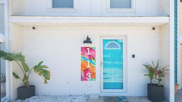 A white brick building with a blue door, a colorful vertical sign, and two potted plants.