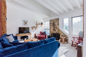A cozy living room with a blue sofa, a red armchair, a stone fireplace, and a television on a wooden stand. - Updated 3Br/3Ba with Panoramic Views. Only Steps to Canyon Lodge! (Mammoth Lakes)