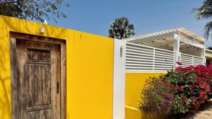 A yellow building with a wooden door, a white pergola, and a planter with red flowers.
