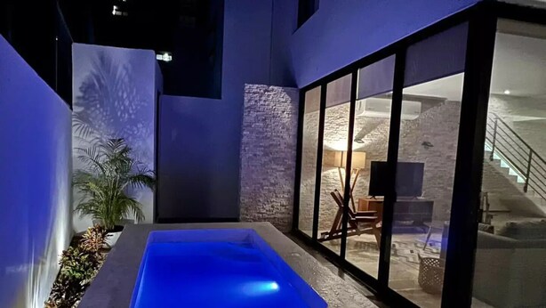 A modern outdoor pool area with a blue pool, stone walls, and a view of the interior through large glass doors. - Casa Montes de Amé, gran ubicación y comodidad (Mérida)