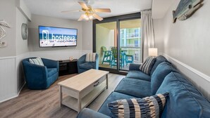 A modern living room with a blue sofa set, a wooden coffee table, and a ceiling fan. A flat-screen TV is mounted on the wall, and there is a sliding glass door leading to an outdoor area with green chairs. - Harbour Place 310 (Orange Beach)