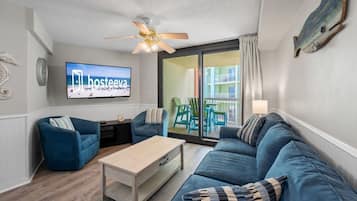 A modern living room with a blue sofa set, a wooden coffee table, and a ceiling fan. A flat-screen TV is mounted on the wall, and there is a sliding glass door leading to an outdoor area with green chairs.