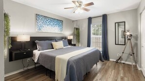 A modern bedroom with a bed, bedside tables, a ceiling fan, a large window with curtains, and a tripod lamp.
