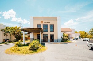 Edifício VillaOeste com entrada coberta, palmeiras e estacionamento.