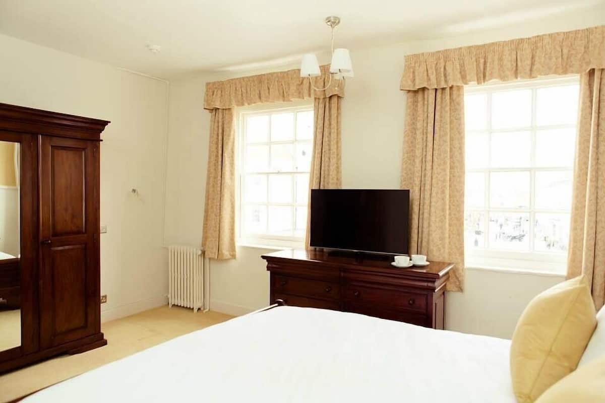 A bedroom with a bed, a TV on a dresser, a wardrobe, and two windows with curtains.
