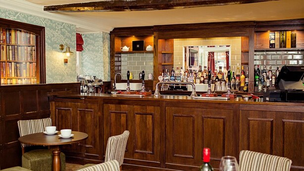 A bar area with a wooden counter, bookshelves, and a seating area with chairs and a table. - Kings Arms Bicester (Bicester)