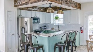 Une cuisine avec des meubles blancs, un îlot central doté de tabourets, et un plafond au style rustique.