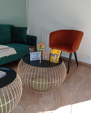 Ein modernes Wohnzimmer mit einem grünen Sofa, einem runden Couchtisch mit Drahtgestell, einem roten Sessel und einem kleinen Tisch mit Snacks und einem Willkommensschild.