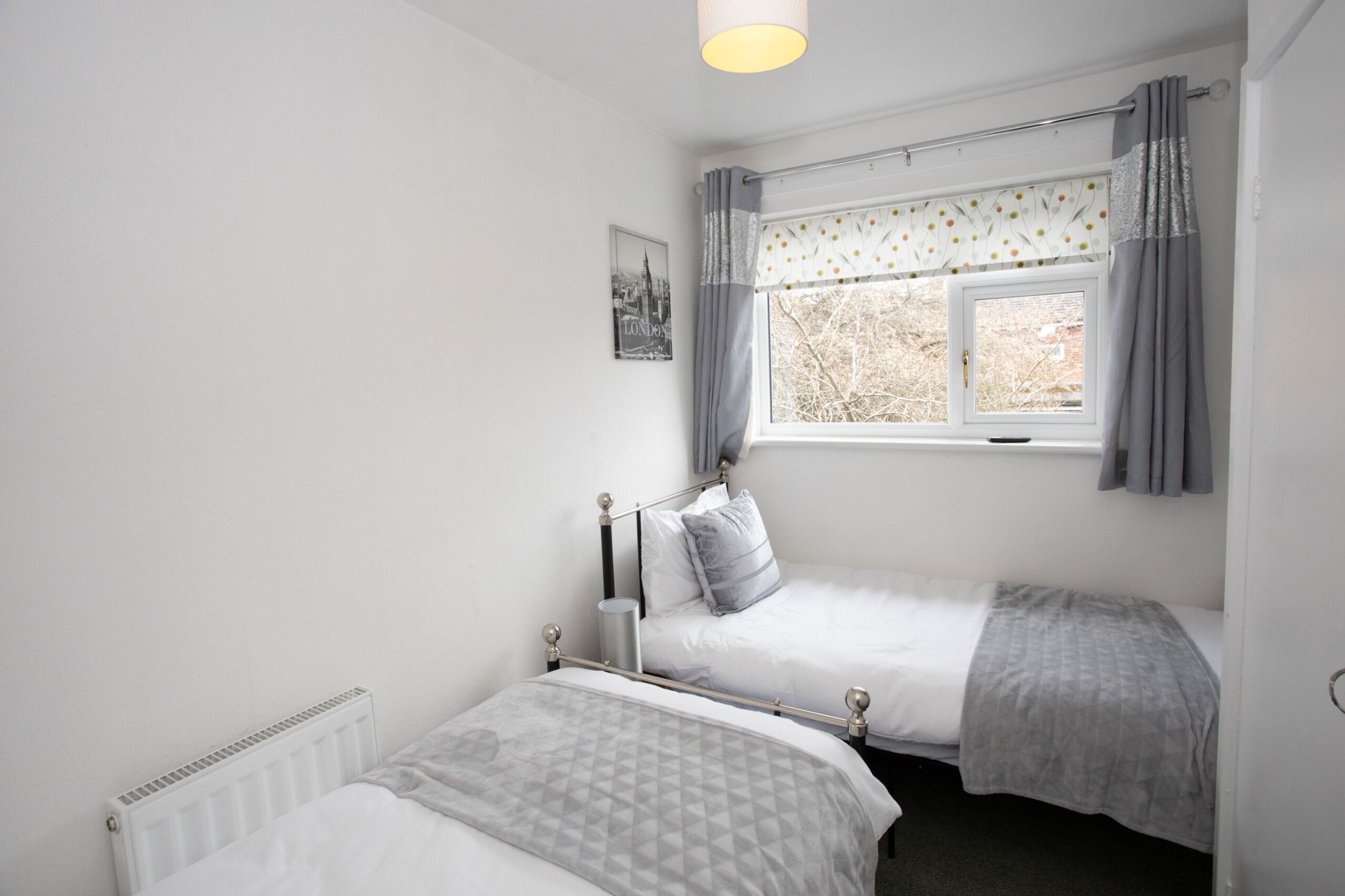A bedroom with two beds, a window with curtains, a radiator, and a wall-mounted picture.