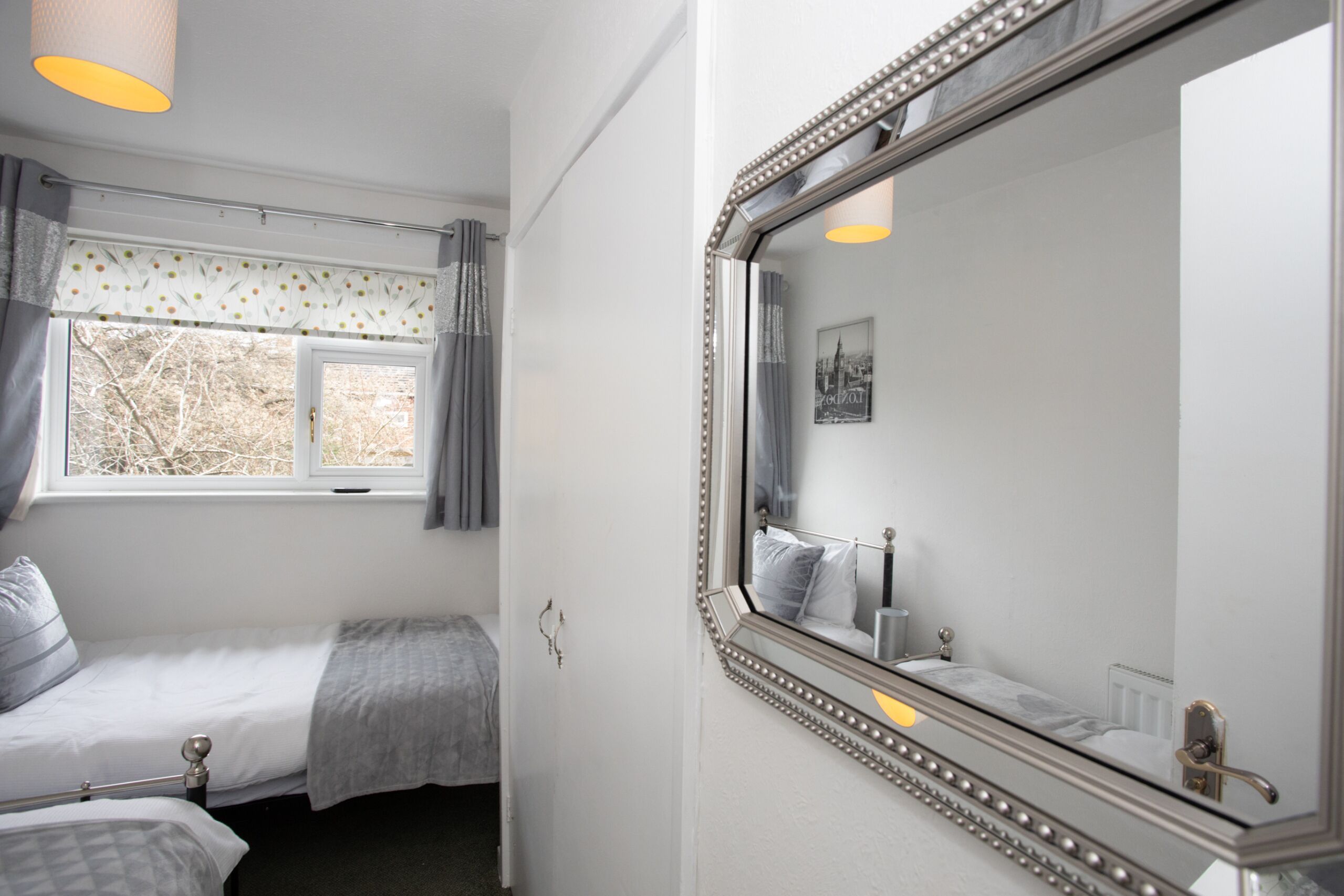 A bedroom with two beds, a window with curtains, a mirror with a decorative frame, and a wall-mounted radiator.
