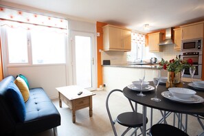 A modern kitchen with wooden cabinets, a dining table set for two, and a sofa with cushions. - Hurley House (Cheadle)