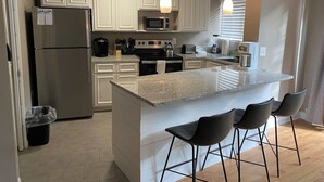 Una cocina moderna con armarios blancos, electrodomésticos de acero inoxidable, encimera de granito y taburetes de bar.