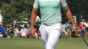 Un campo de golf con un jugador, vestido con una camisa verde clara y blanca a rayas y pantalones blancos, caminando sobre el green.