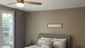 Un dormitorio con cama, ventilador de techo, mesita de noche y una ventana con cortinas, además de un cartel en la pared.