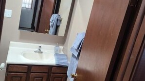 A bathroom with a wooden vanity, a sink, a mirror, and a towel hanging on the door. - Comfy bungalow (Carthage)