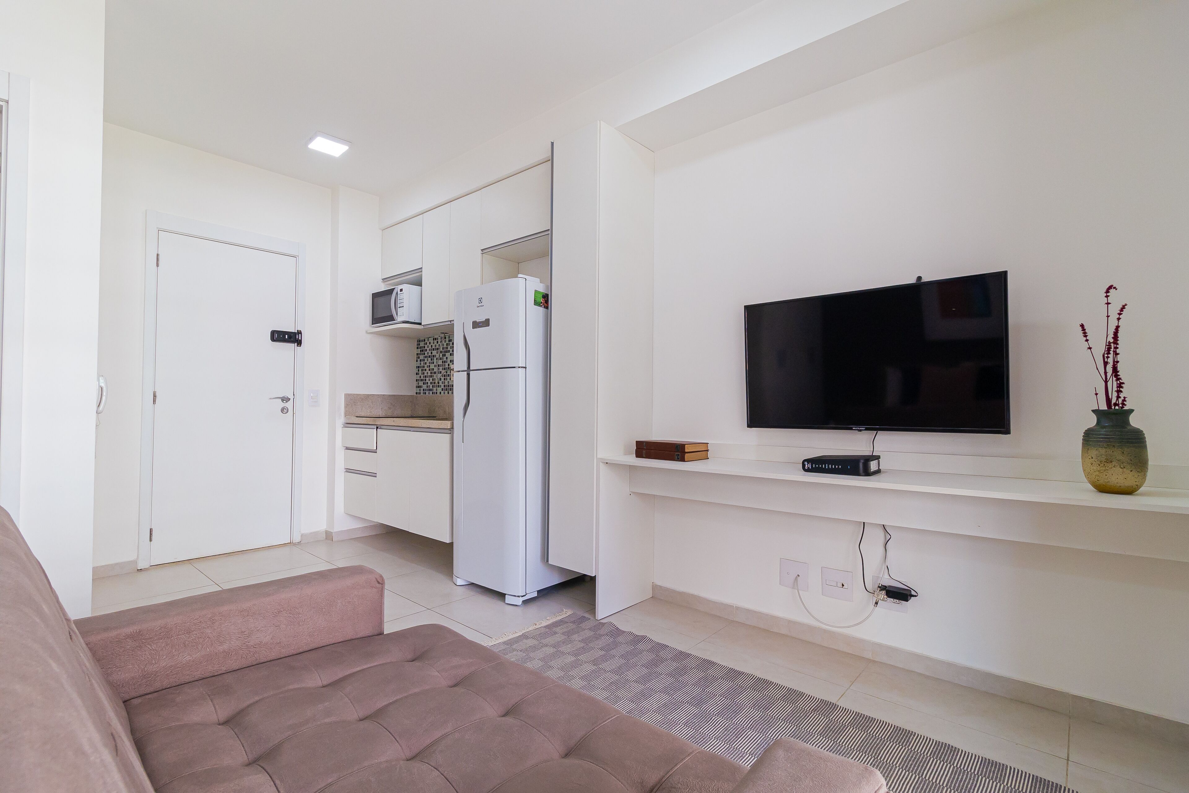 A modern living room with a sofa, a flat-screen TV mounted on the wall, a kitchen area with a refrigerator and microwave, and a door with a handle.
