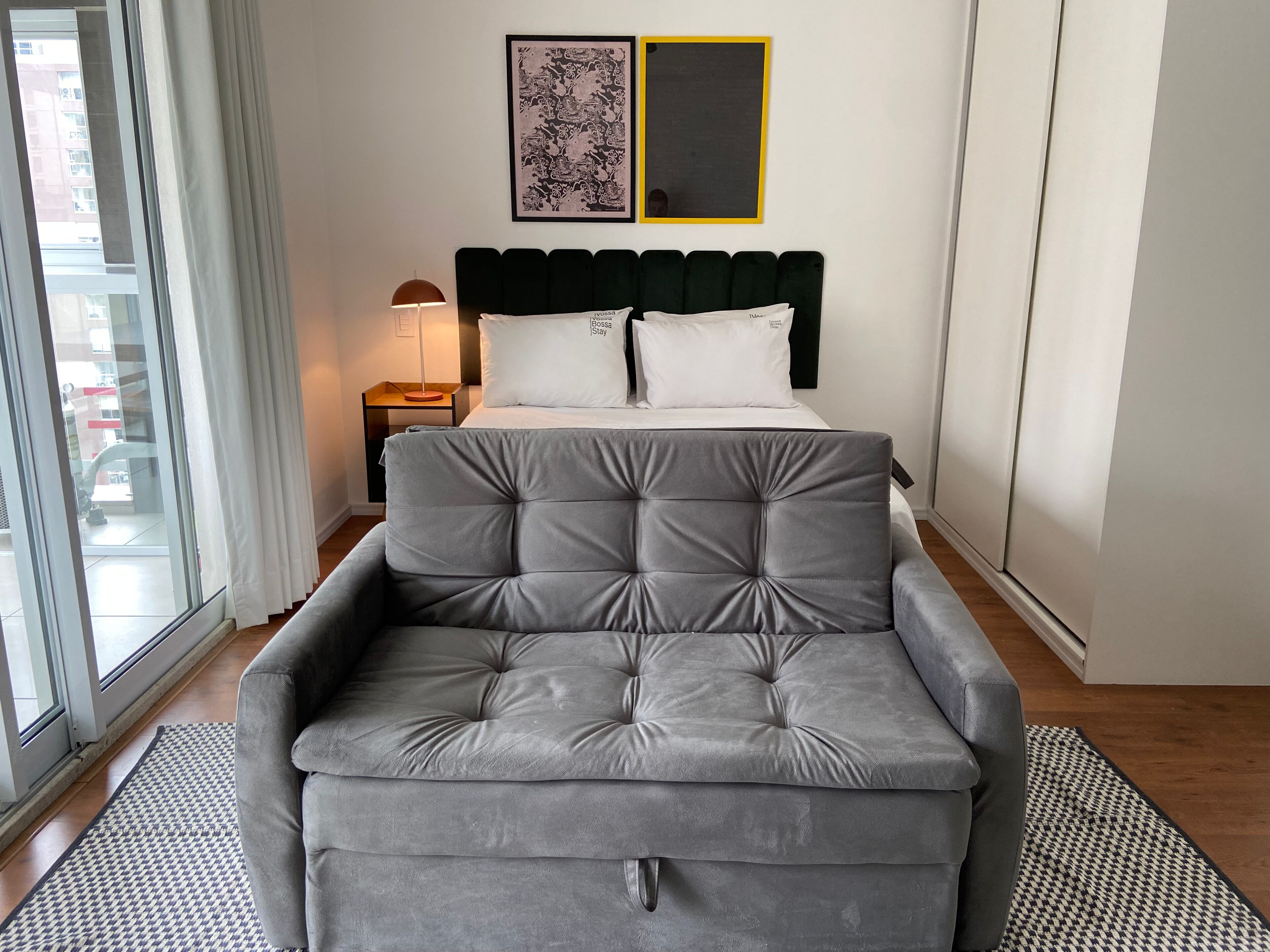 A modern bedroom with a grey sofa, a bed with white bedding, a nightstand with a lamp, and a large window.
