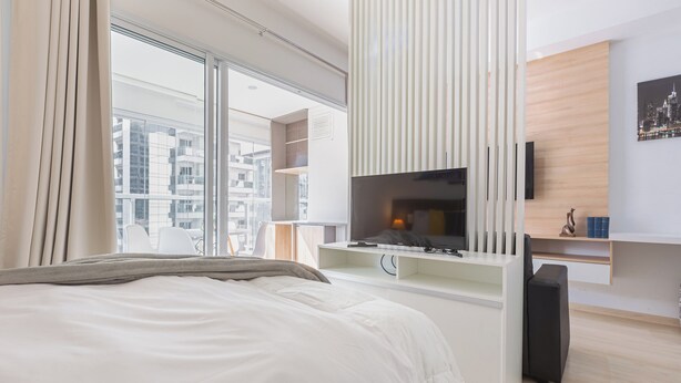 Una habitación de hotel moderna con una cama grande, un televisor montado en un panel de madera, un escritorio con una silla y vistas a la ciudad a través de amplias ventanas.