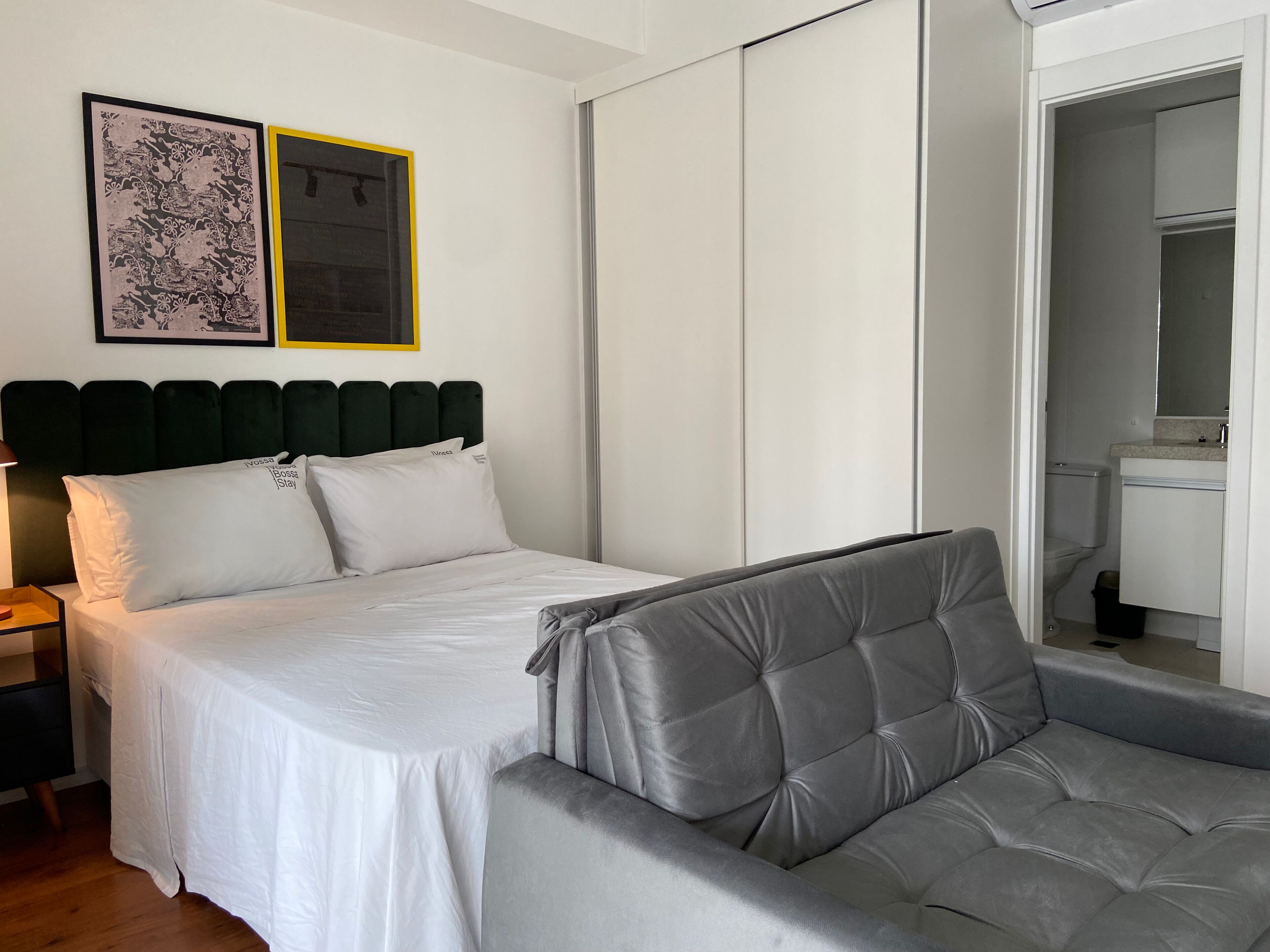 A neatly arranged bedroom with a bed, a grey sofa, a wardrobe, and a bathroom visible through an open door.