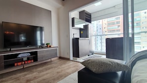 Una sala moderna con un televisor de pantalla plana, un mueble empotrado para entretenimiento y un ventanal corredizo que da a un balcón con vistas a la ciudad.