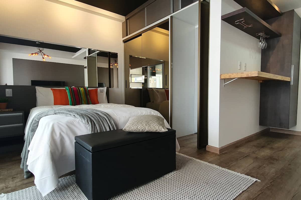 A modern bedroom with a large bed, bedside tables, a bench, a mirror, and a built-in wardrobe.