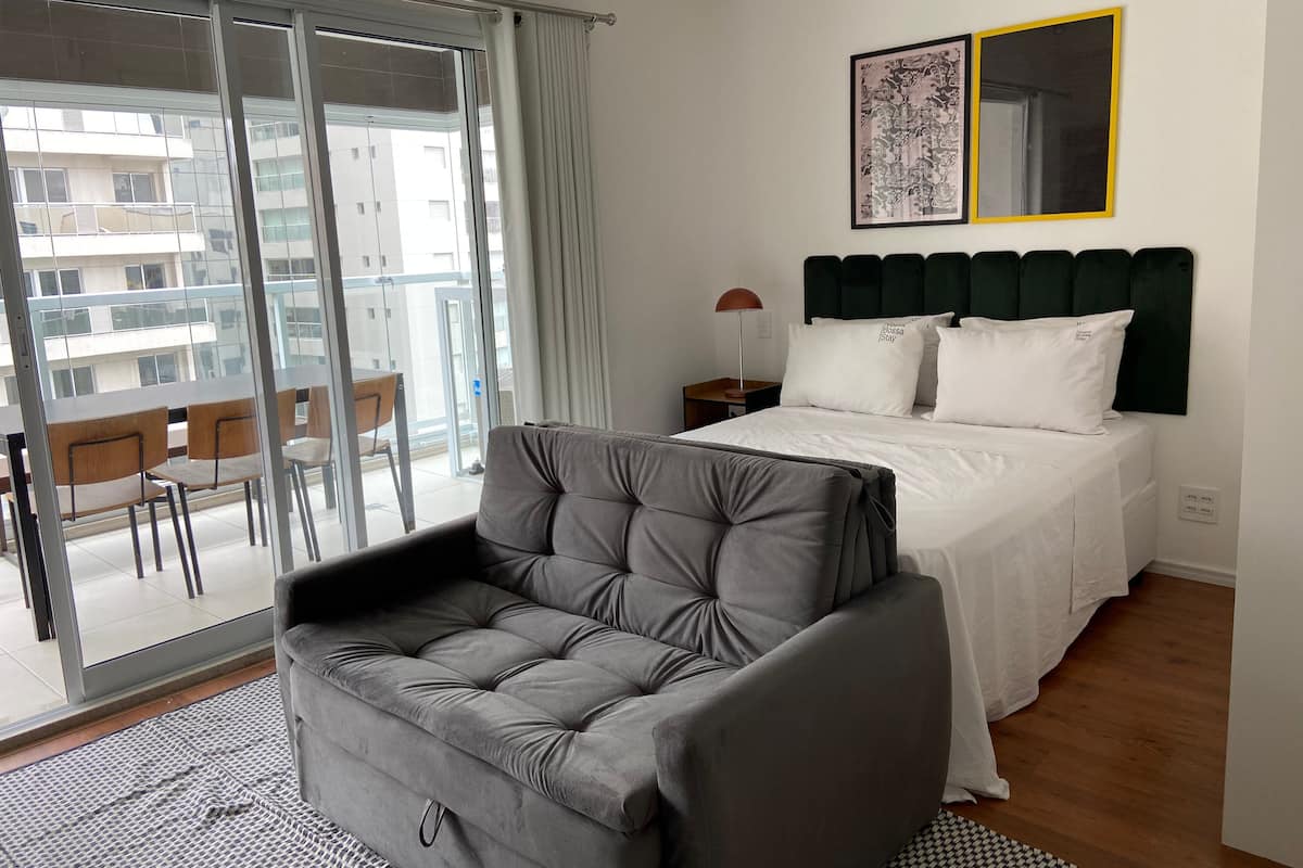 A modern hotel room with a bed, a grey sofa, a dining area with chairs, and a large window with a view of the city.