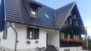 A two-story house with a steeply pitched roof, a stone foundation, and a front porch with a table and chairs. - Soba Deluxe s king size krevetom (Plitvica Selo)