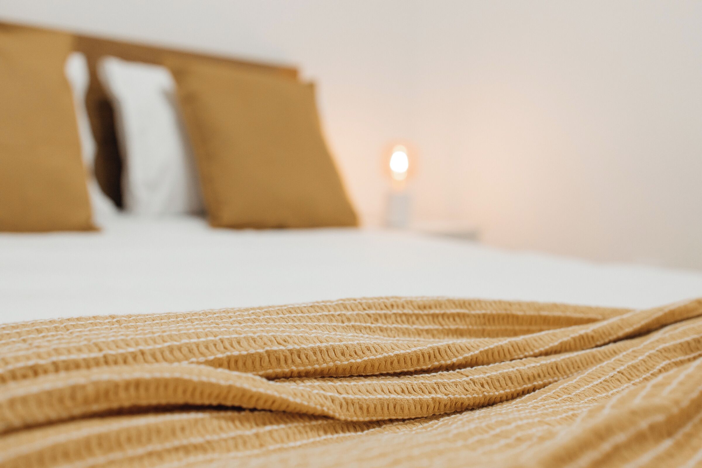 A bed with a textured blanket and pillows, a bedside table with a lamp, and a window with a view of the sky.