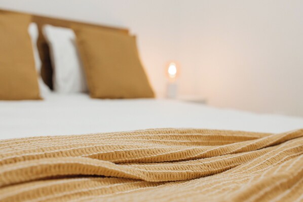 A bed with a textured blanket and pillows, a bedside table with a lamp, and a window with a view of the sky.