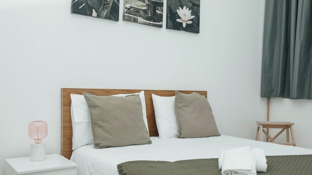 A neatly made bed with a green throw blanket, a bedside table with a pink lamp, and a small wooden stool.