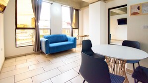 Una habitación con un sofá azul, una mesa redonda con sillas y ventanas amplias con cortinas.