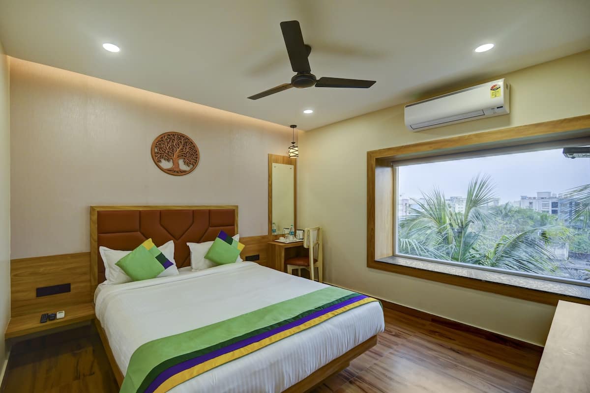 Una habitación de hotel con cama, ventilador de techo, unidad de aire acondicionado, ventana con vistas y mesita de noche de madera.