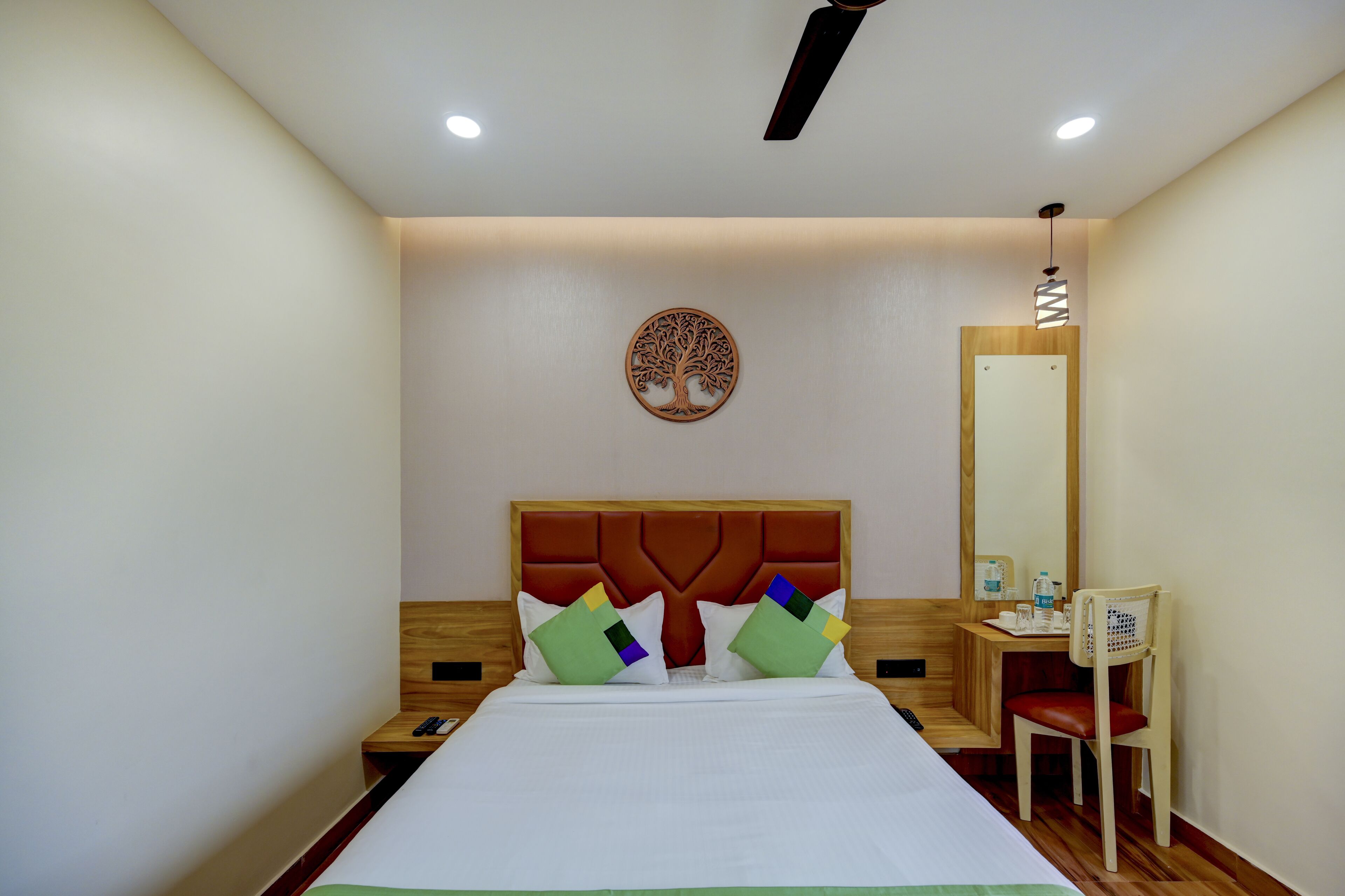 A hotel room with a bed, a wooden nightstand, a chair, a desk, and a mirror.
